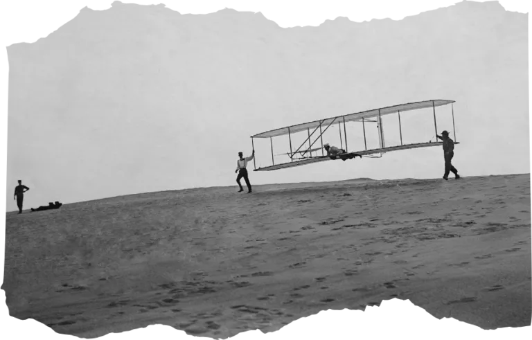 Wright Brothers' glider iwth a single vertical rudder before its flight, Oct. 10, 1902. Orville lies prone in glider as Wilbur and Dan Tate hold the wing tips at left and right. Kitty Hawk, North Carolina.
