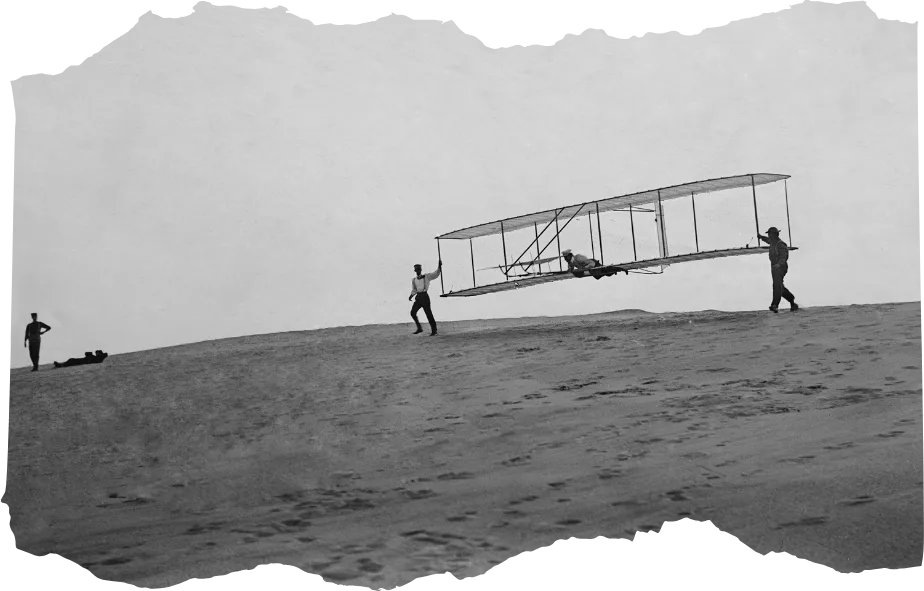 Wright Brothers' glider iwth a single vertical rudder before its flight, Oct. 10, 1902. Orville lies prone in glider as Wilbur and Dan Tate hold the wing tips at left and right. Kitty Hawk, North Carolina.