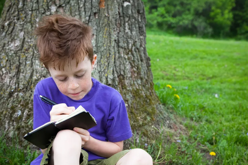 boy in purple shirt with journal, writing outside under a tree